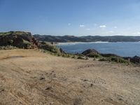 a dirt road runs along the coast, with hills in the background and a blue sky above