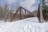 an abandoned wooden and rusted foot bridge in the snow with trees around it on a cloudy day