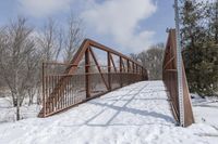 an abandoned wooden and rusted foot bridge in the snow with trees around it on a cloudy day