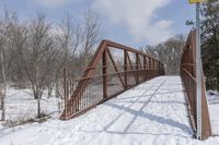 an abandoned wooden and rusted foot bridge in the snow with trees around it on a cloudy day