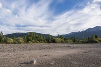a dirt field and rocks sitting under clouds above the ground with trees on both sides