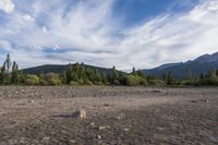 a dirt field and rocks sitting under clouds above the ground with trees on both sides