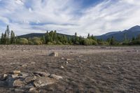 a dirt field and rocks sitting under clouds above the ground with trees on both sides