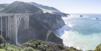 an elevated bridge is extending into the ocean near cliffs and cliffs in the background with waves crashing onto the water