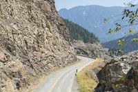 a motorcycle rider rides down a mountain road next to rocky cliffs and trees as seen from a distance