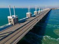 a bridge over the ocean with construction equipment in the background and two people walking on it