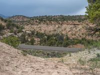 a dirt mountain and dirt road with bushes, rocks, shrubs, and clouds in the sky