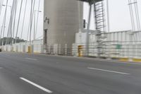 a gray car driving over a bridge with lights on in the distance, the bridge and trees near