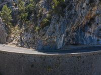 the motorcycle is riding down the road by a huge rock wall and steeply slope