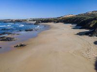 a sandy beach with a little ocean next to it, next to a hill, with a building on the far shore