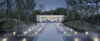 a fountain and the mountains are seen in this photo from behind it, along with lights, on a white walkway, surrounded by trees