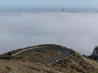 an image of clouds and city below the road in the hills above it, with a car traveling on one