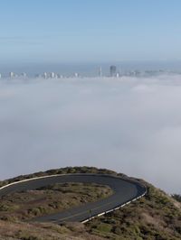 an image of clouds and city below the road in the hills above it, with a car traveling on one