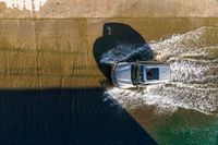 an overhead view of a white car driving down the road near the water's edge