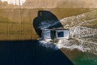 an overhead view of a white car driving down the road near the water's edge