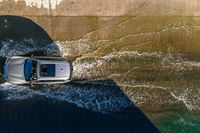 an overhead view of a white car driving down the road near the water's edge