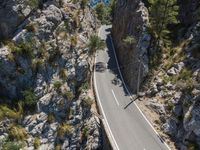 the road is winding between the cliffs and trees, with two motorcycles on it in mid distance