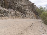 a motorcycle rides on an empty dirt road with large rocks and shrubs near it as another drives down it