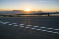 the road is empty and the sun rises behind some mountains along the highway side, with one half in the distance