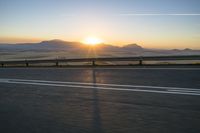 the road is empty and the sun rises behind some mountains along the highway side, with one half in the distance