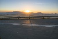 the road is empty and the sun rises behind some mountains along the highway side, with one half in the distance