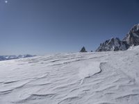 snow covered ground near rocks in the middle of mountain range during wintertime under a sunny blue sky