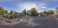 360 - lens photo of bicycles parked on brick path along canal side of street area in urban area with trees and building in background