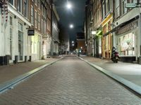 a narrow brick paved city street in the evening, with a man sitting on a motorcycle parked next to it