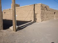 a lone telephone pole is casting a shadow on the stone wall of an adobe hut