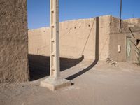 a lone telephone pole is casting a shadow on the stone wall of an adobe hut