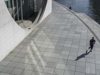 a group of people walking along a walkway next to water and buildings with windows and shadows on it