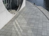 a group of people walking along a walkway next to water and buildings with windows and shadows on it