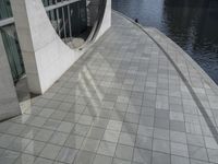 a group of people walking along a walkway next to water and buildings with windows and shadows on it