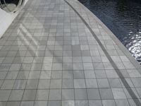 a group of people walking along a walkway next to water and buildings with windows and shadows on it