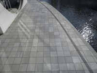 a group of people walking along a walkway next to water and buildings with windows and shadows on it