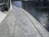 a group of people walking along a walkway next to water and buildings with windows and shadows on it