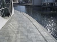 a group of people walking along a walkway next to water and buildings with windows and shadows on it