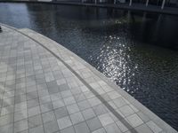 a group of people walking along a walkway next to water and buildings with windows and shadows on it