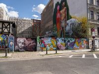 a wall with different colorful artwork on it in a city with buildings around and the street has a bike path going under it