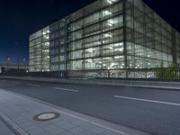 the large building has glass and lights on it's sides on the sidewalk beside a paved roadway