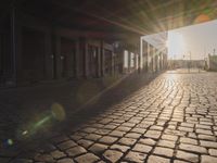 the sun shines behind a row of buildings along a street that has a brick sidewalk and two buildings along side it