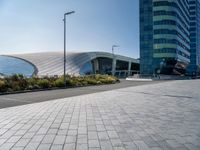 the side of a building that is made of glass and has a circular roof that overlooks an outdoor area with a paved pathway in front of vegetation to it