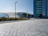 the side of a building that is made of glass and has a circular roof that overlooks an outdoor area with a paved pathway in front of vegetation to it