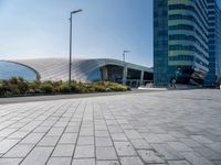 the side of a building that is made of glass and has a circular roof that overlooks an outdoor area with a paved pathway in front of vegetation to it