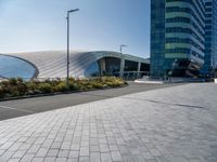 the side of a building that is made of glass and has a circular roof that overlooks an outdoor area with a paved pathway in front of vegetation to it