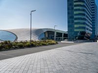 the side of a building that is made of glass and has a circular roof that overlooks an outdoor area with a paved pathway in front of vegetation to it