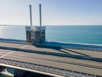 a bridge over the ocean with construction equipment in the background and two people walking on it