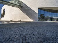 a circular stone structure with a glass facade and two benches under it on a brick walkway near a modern glass building