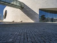 a circular stone structure with a glass facade and two benches under it on a brick walkway near a modern glass building