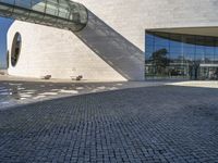 a circular stone structure with a glass facade and two benches under it on a brick walkway near a modern glass building